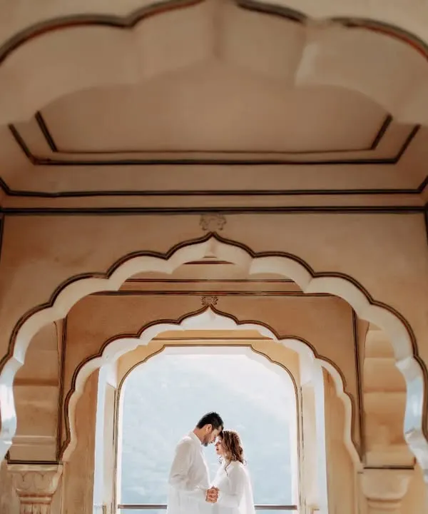A beautifully symmetric shot using the layered arches of a Jaipur palace to frame the couple. This intimate forehead-to-forehead pose creates a sense of unity and peace amidst the grandeur.