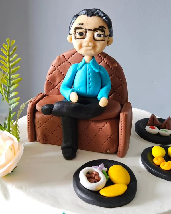 I believe the best cakes tell a story. For this special retirement cake, I handcrafted an edible figurine of the client's father relaxing in his favorite armchair, surrounded by miniature versions of his favorite foods. Every detail, from the quilted texture of the fondant sofa to the tiny edible mangoes, is made by hand to create a truly personal and memorable centerpiece.