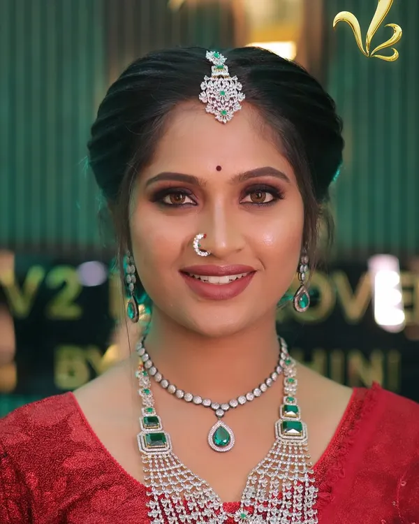 A full view of a stunning modern reception look. The bride is styled in a vibrant red saree, paired with a multi-layered diamond and emerald necklace set. The makeup is glamorous yet fresh, creating a sophisticated look perfect for an evening celebration.