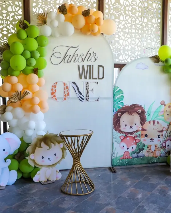 A 'Wild One' jungle theme decoration for a first birthday party. This setup includes a custom backdrop, organic balloon garlands in green, orange, and white, and cute animal cutouts like a lion and elephant to complete the safari look.