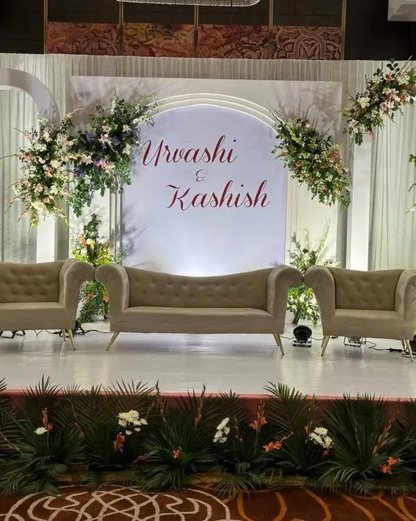 A chic and modern engagement stage design. It features a clean white backdrop with arched panels, the couple's names in elegant calligraphy, and asymmetrical floral arrangements for a sophisticated look.