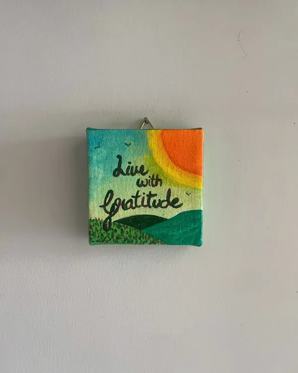 "Live with Gratitude." This mini 4x4 inch canvas is a simple, colorful reminder to appreciate the small joys, featuring a bright sun over green hills.
