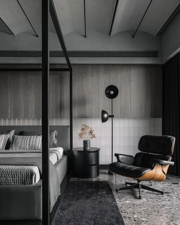 The master bedroom in the Vaulted Haus features a large four-poster bed and an Eames lounge chair. The headboard wall combines backlit frosted glass bricks with grey veneer strips, creating a multi-layered, tactile surface.