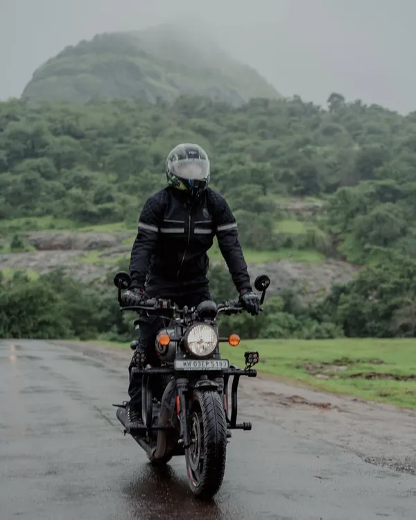 A rider in full gear navigates a wet, winding road on a Royal Enfield during the monsoon. The lush green hills of the Western Ghats in the background create a classic road trip scene.