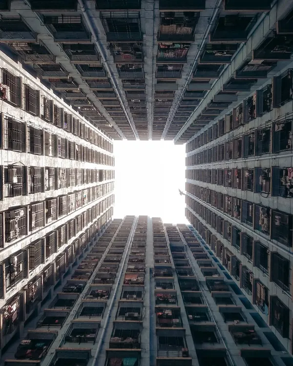 A worm's-eye view looking straight up at the sky, perfectly framed by the repeating patterns of a dense residential building. This shot creates a feeling of being enclosed by the city's vertical growth, a classic Hong Kong-style "monster building" perspective.