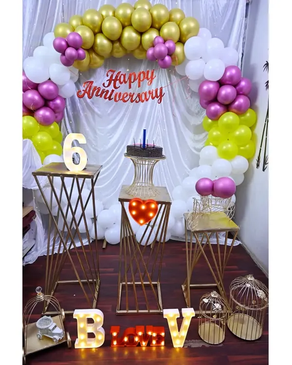 An anniversary party setup with a white, gold, and pink balloon arch. The decoration includes modern gold pedestals, birdcage props, and marquee letters spelling out initials.