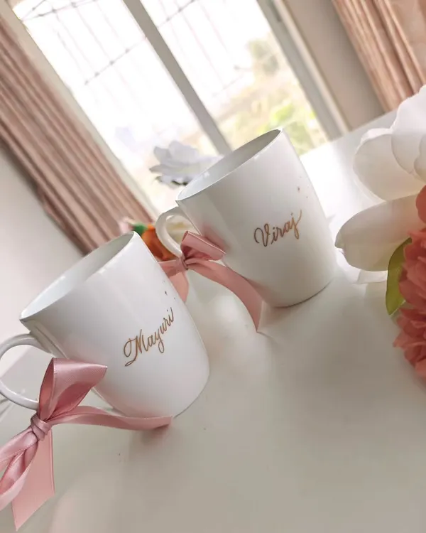 A lovely anniversary gift for a couple. These white ceramic mugs are personalized with the names 'Mayuri' and 'Viraj' in an elegant gold script, tied with pink ribbons for a celebratory finish.