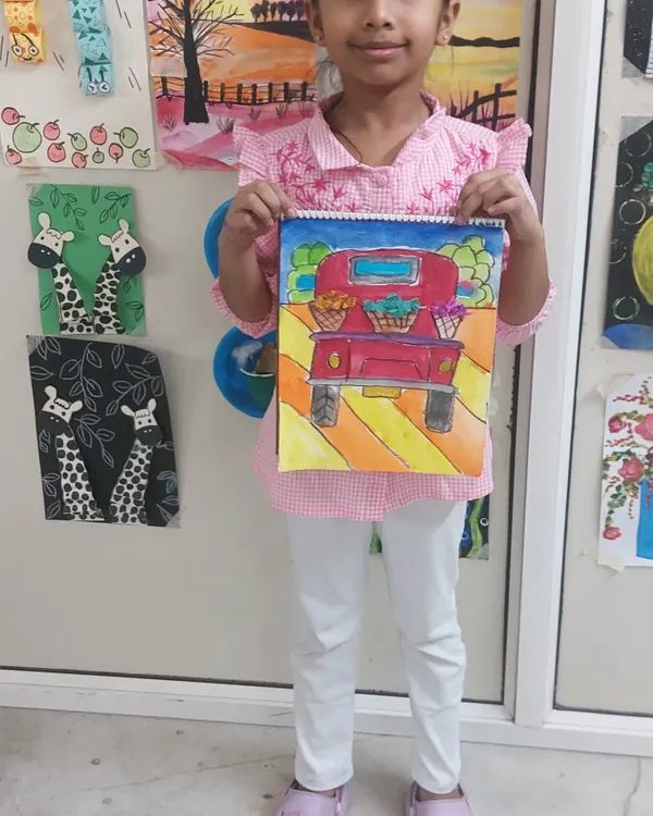 A proud student displays her colorful truck painting. Our art classes guide children through different techniques like watercolor and pattern drawing to create vibrant, detailed artwork.