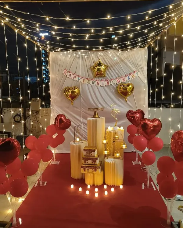 I create magical, intimate anniversary surprises right in your home. This romantic canopy setup features a curtain of warm fairy lights, a classic 'Happy Anniversary' banner, and elegant red heart balloons to create a truly memorable and cozy atmosphere.
