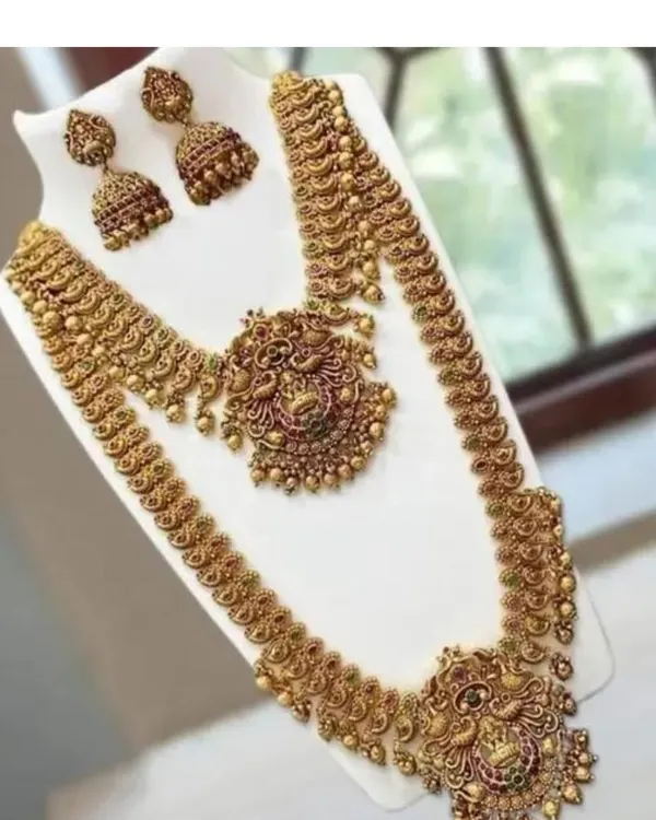 A complete temple jewellery set in antique gold finish, laid out on a display. This set includes a short necklace, a long haram, and matching jhumkas, all featuring intricate peacock and goddess motifs.