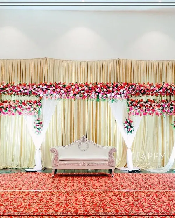 An elegant and budget-friendly stage decoration featuring cream-colored drapes and dense runners of red and pink flowers. This clean and classic design provides a beautiful backdrop without overwhelming the space.
