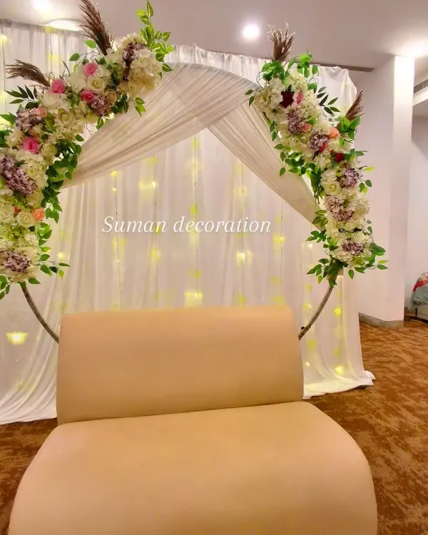 This backdrop features a circular arch decorated with artificial flowers and pampas grass. The white fabric drapes and fairy lights create a soft, glowing background.