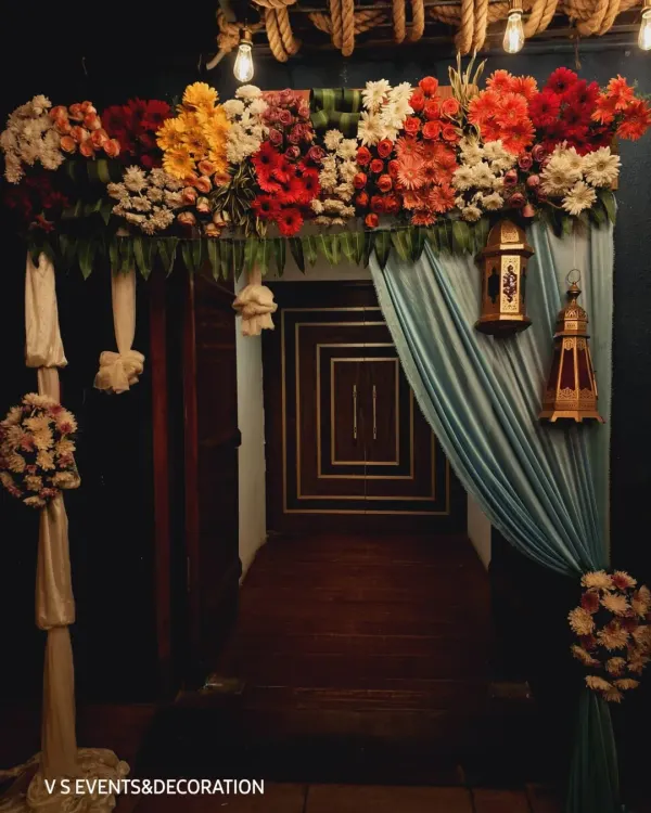An entrance arch designed for an evening engagement. The decor features a thick arrangement of red, orange, and white flowers, with draped fabric and hanging Moroccan lanterns to create a warm and inviting entryway.