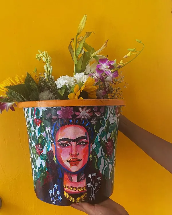 This hand-painted planter features a portrait of Frida Kahlo, surrounded by a lush floral pattern. It's a functional piece of art designed to bring life and color to any room or balcony.