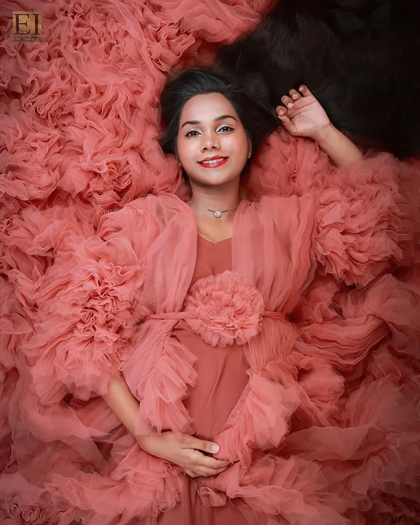 A moment of pure serenity. This artistic overhead shot captures the mom-to-be enveloped in a magnificent ruffled pink gown, creating a visual of her blooming like a flower. This is one of our signature studio portraits.