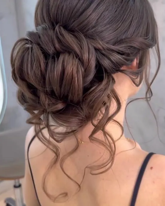 An intricate and voluminous low bun, perfect for a bride or bridesmaid. The soft tendrils framing the face add a touch of romance to this classic and elegant updo.