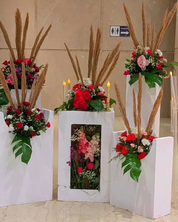 These modern floral arrangements combine red roses and pampas grass in geometric white stands. The central stand features a unique shadow box design, adding an artistic touch.