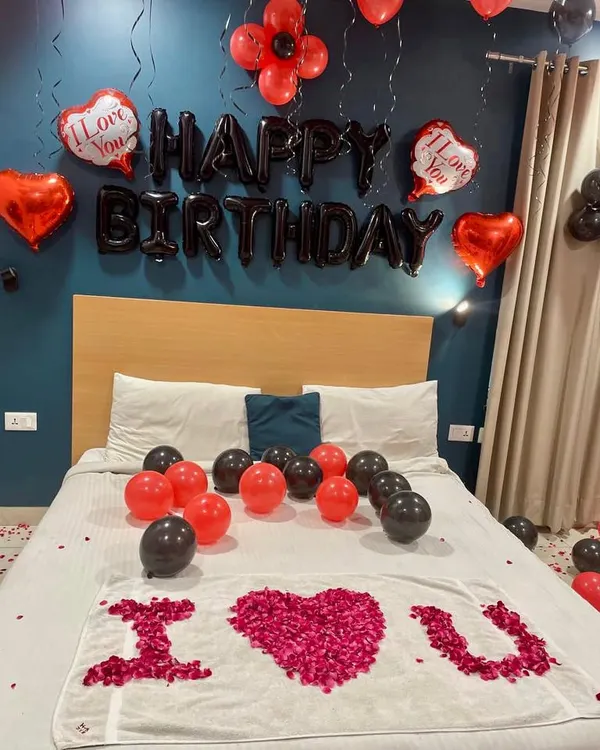 A classic romantic birthday room decoration. This setup includes "Happy Birthday" foil balloons, red and black latex balloons, and a heart-shaped "I Love You" message created with rose petals on the bed.