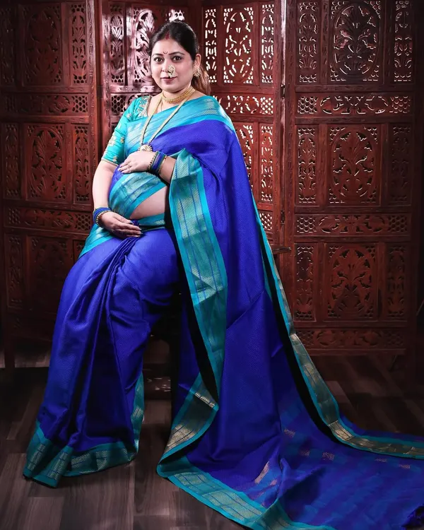A regal portrait of a mother-to-be in a stunning blue and turquoise silk saree. The traditional jewelry and pose against a carved wooden screen create a timeless and culturally rich image.