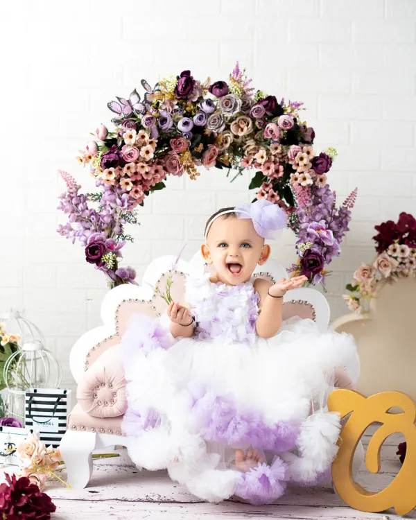 A beautiful portrait for a first birthday celebration. This little girl is all smiles in her fluffy purple and white dress, sitting on a miniature couch under a floral arch.