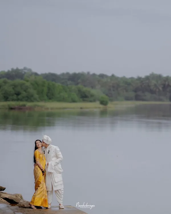 A groom-to-be gently kisses his partner's cheek as they stand by a calm, expansive river. This is a perfect example of a tender, romantic moment captured in a serene natural setting.