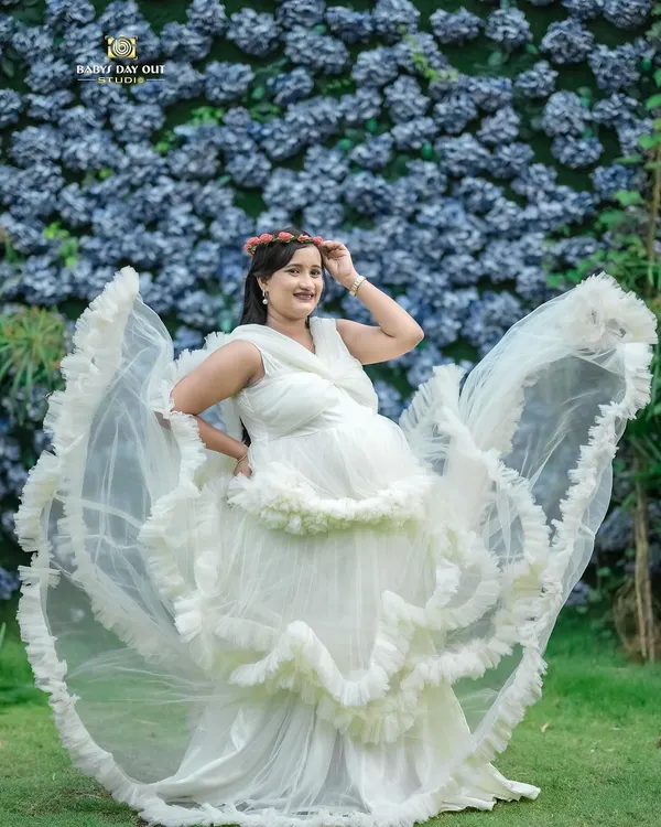 A full-length view of our popular white ruffled gown against a beautiful wall of blue flowers. This outdoor setup at our studio is perfect for creating dramatic and angelic maternity portraits.