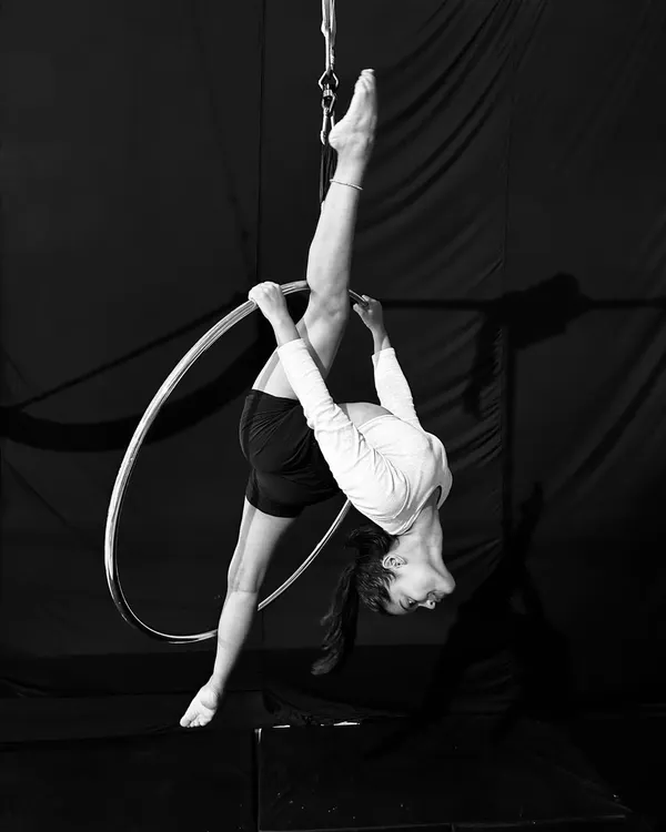 This stunning black and white photo captures an advanced student holding a perfect vertical split on the aerial hoop, showcasing incredible flexibility and control.
