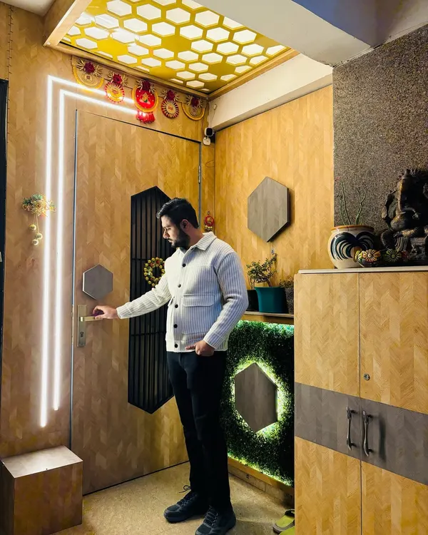 A stunning main entrance at Prateek Laurel. The door is flanked by vertical LED profile lights, and the foyer features a honeycomb-patterned ceiling and a shoe cabinet with a green moss accent.