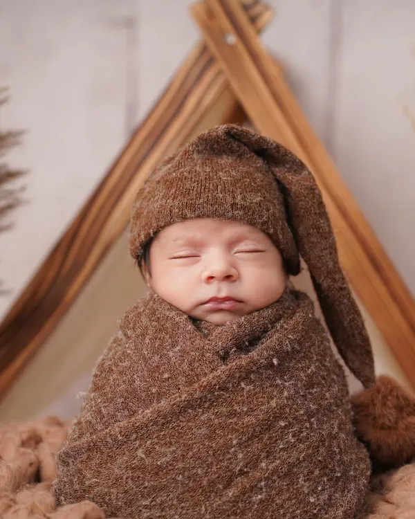 A peaceful newborn baby is swaddled in a cozy brown wrap and matching pom-pom hat, posed in a rustic, camping-themed setup with a wooden tent frame.