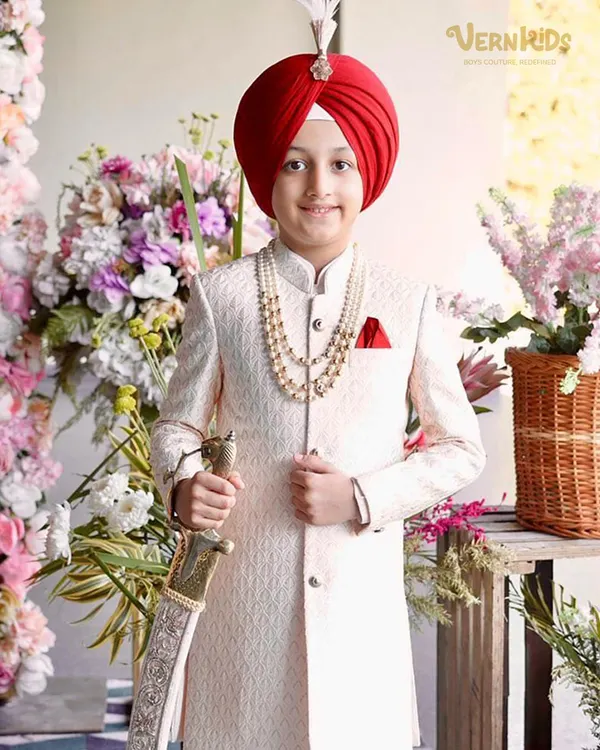This little star is ready for his big moment in a stunning off-white sherwani, complete with a pearl necklace and a royal red turban. It is a timeless look, tailored for unforgettable wedding celebrations.