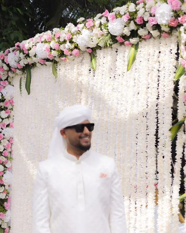 The groom looking cool and confident in his all-white Nikaah outfit, complete with sunglasses. A stylish portrait against a beautiful floral backdrop.