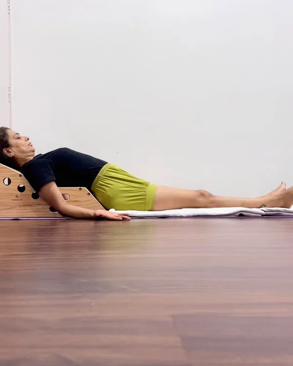 This is a supported Savasana on a Simhasana Box, a prop designed to open the chest and support the spine's natural curves. This posture encourages deeper breathing and helps the body shift into a state of rest and recovery.