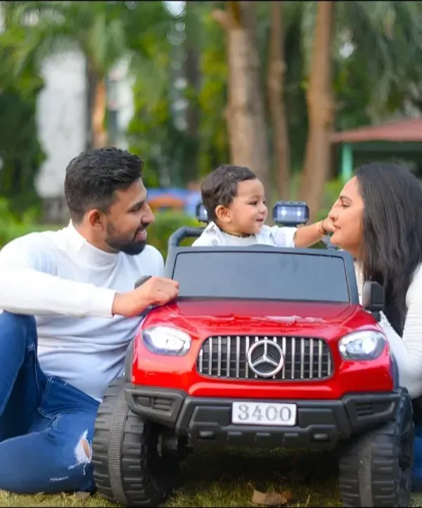 A candid moment from a family photoshoot in the park. The parents share a loving moment with their child, who is having a blast in his toy car.