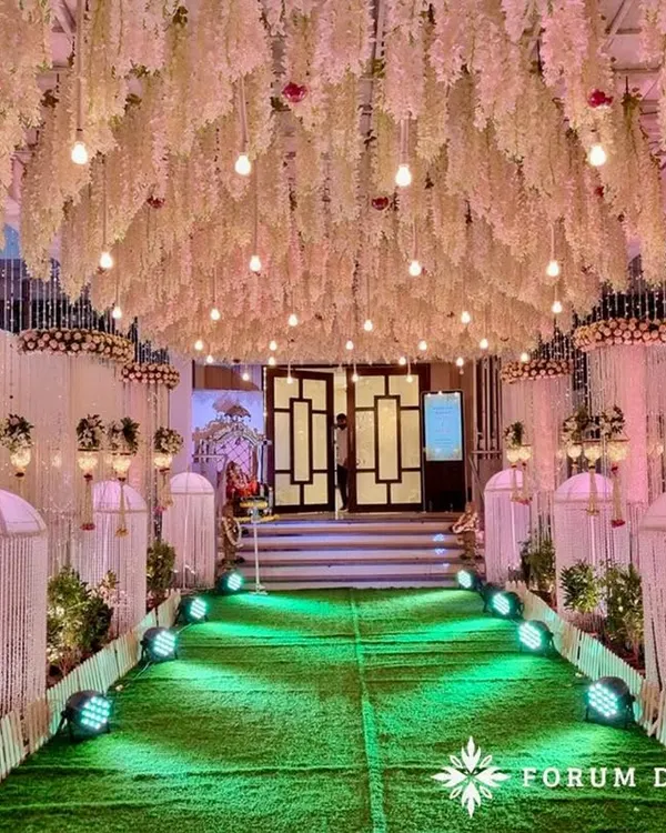 This dreamy pastel walkway creates a magical path for guests. The ceiling is a dense canopy of hanging white wisteria and light bulbs, creating a starry night effect. The green turf path is lined with draped pillars and traditional hangings, making the walk to the main event an experience in itself.