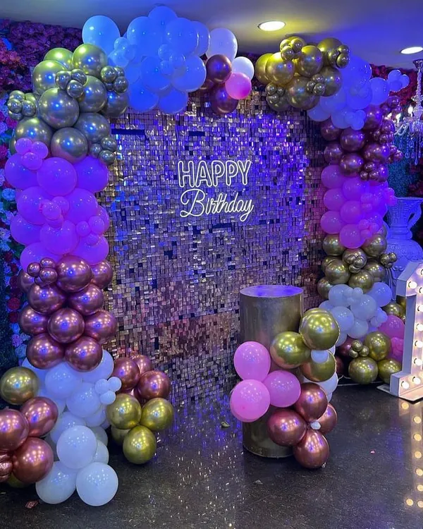 A glamorous birthday setup with a gold shimmer wall as the centerpiece. The decor is accented with purple, pink, and gold balloons, a 'Happy Birthday' neon sign, and a light-up number '1'.