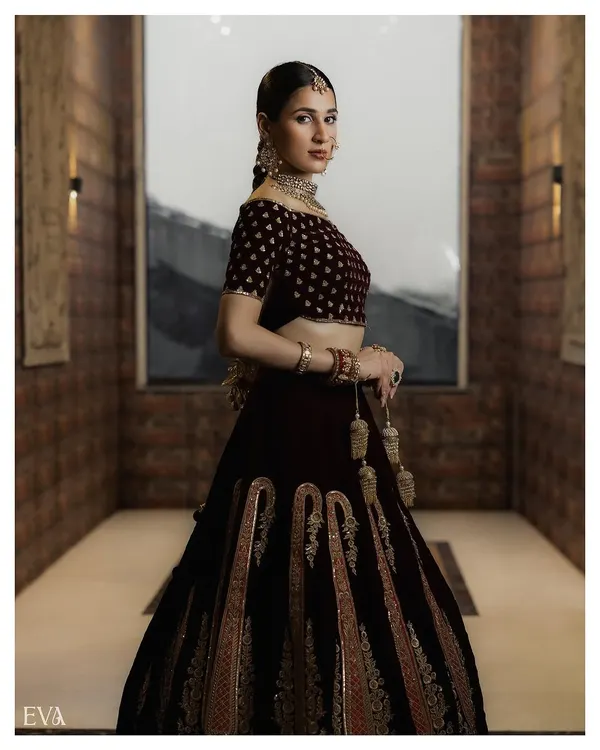 A full-length portrait showcasing a magnificent velvet lehenga with detailed embroidery. This shot captures the entire silhouette and the grandeur of the bridal outfit within a well-composed architectural setting.