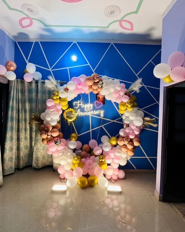 This indoor anniversary decoration features a full circular balloon arch in pastel pink, white, and gold tones. A 'Happy Anniversary' neon sign hangs in the center, creating a beautiful focal point in the room.