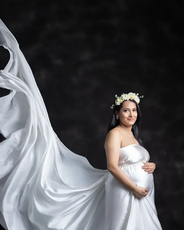 A solo portrait capturing the pure elegance of motherhood. This shot features one of our flowing white satin gowns with a dramatic fabric toss against a classic dark backdrop, creating a timeless and angelic look for the mom-to-be.