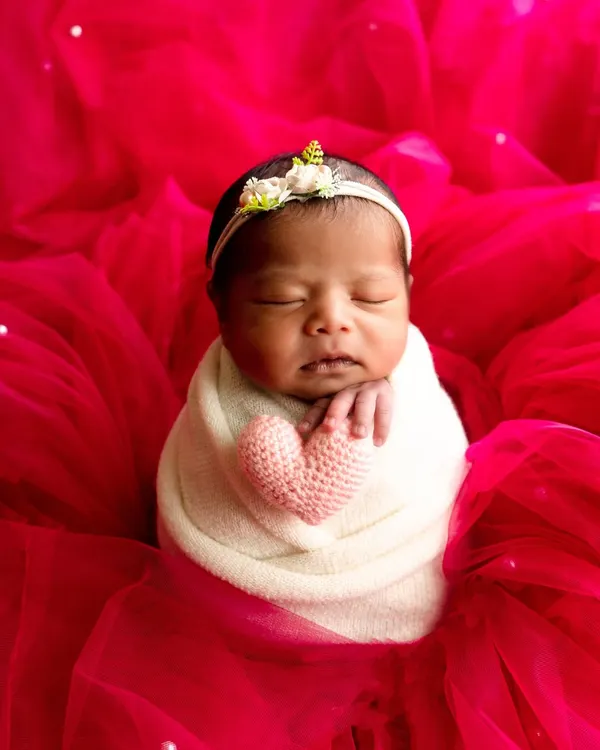 A classic newborn portrait of a baby girl swaddled in white, holding a pink knit heart against a vibrant red tulle background.