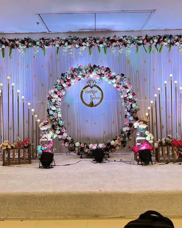 A breathtaking reception stage setup in a grand hall, featuring a large circular floral arch made of pink, white, and blue flowers. The backdrop is a soft white curtain, elegantly lit with warm, standing candle-like lights, creating a magical and romantic atmosphere for the perfect wedding celebration.