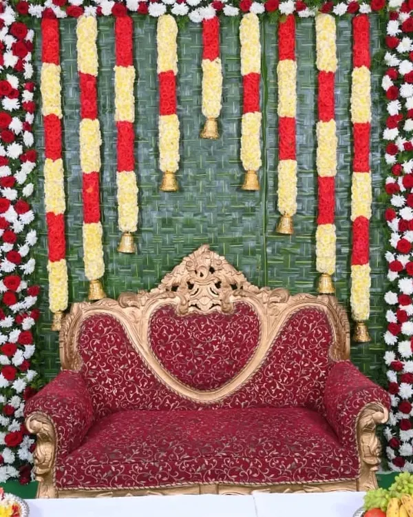 A traditional Seemantha backdrop made with a woven coconut leaf mat. The stage is decorated with red and white floral borders and hanging garlands of yellow chrysanthemums and bells.