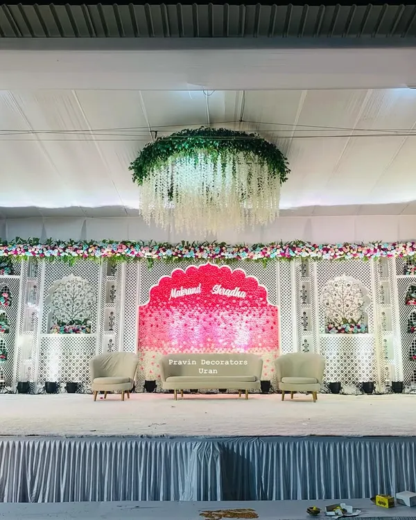 This indoor wedding stage combines royal and floral elements. We used white jali panels, a custom pink backdrop with the couple's names, and a stunning floral chandelier with hanging wisteria to create a rich, layered look.