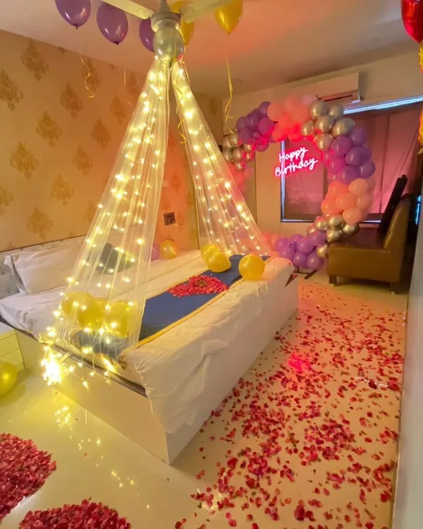 A complete romantic room decoration for a birthday, featuring a fairy light canopy over the bed, a balloon arch, and a floor scattered with rose petals.