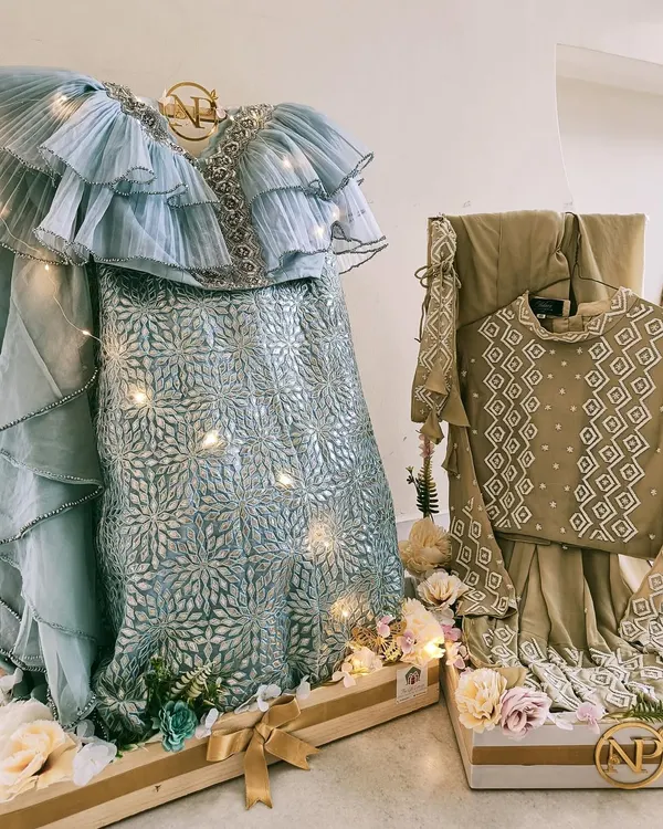 A breathtaking display for a modern bride. This pastel blue ruffled lehenga is presented on a custom wooden stand, adorned with fairy lights and a personalized monogram, creating a stunning focal point for the trousseau.