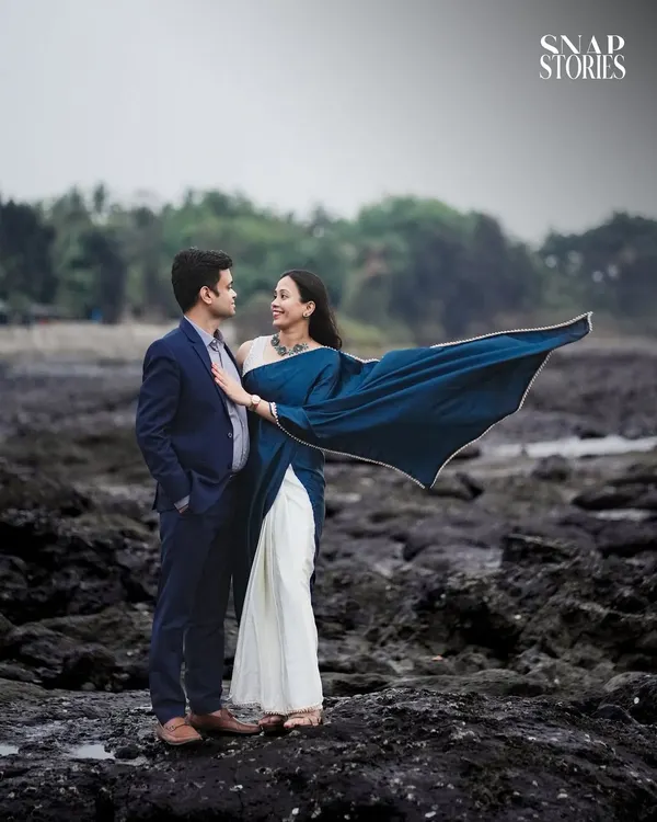 A dramatic shot of a couple on a rocky shore, with the bride's blue saree flowing in the wind. This creates a cinematic and powerful pre-wedding image.