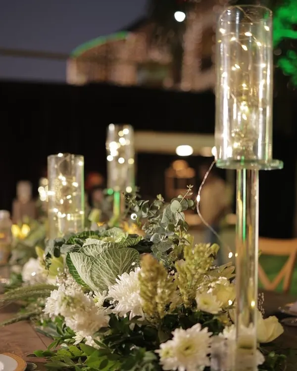 A close-up of a centerpiece featuring fresh white flowers and eucalyptus, with tall glass hurricanes containing fairy lights. This design adds a touch of modern romance and gentle light to the table.