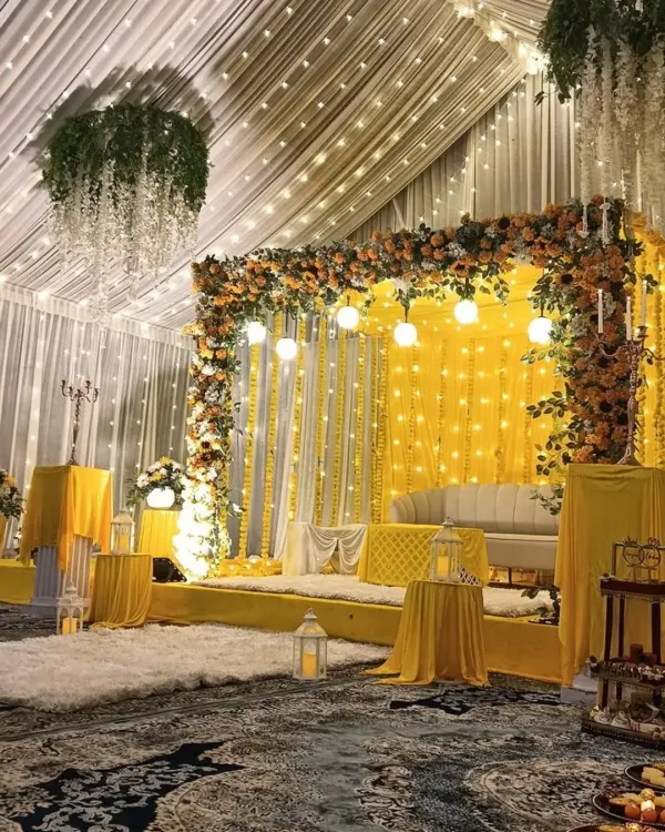 This stage setup for a Haldi ceremony combines a bright yellow backdrop with lush marigold and sunflower arrangements. The hanging lights and white lanterns add a touch of modern elegance to the traditional theme, creating a warm and inviting focal point.