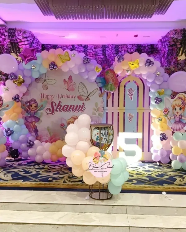 An enchanting fairy-themed stage decoration for a 5th birthday. This setup includes a custom backdrop, pastel balloon arrangements, fairy cutouts, and a large light-up number '5'.