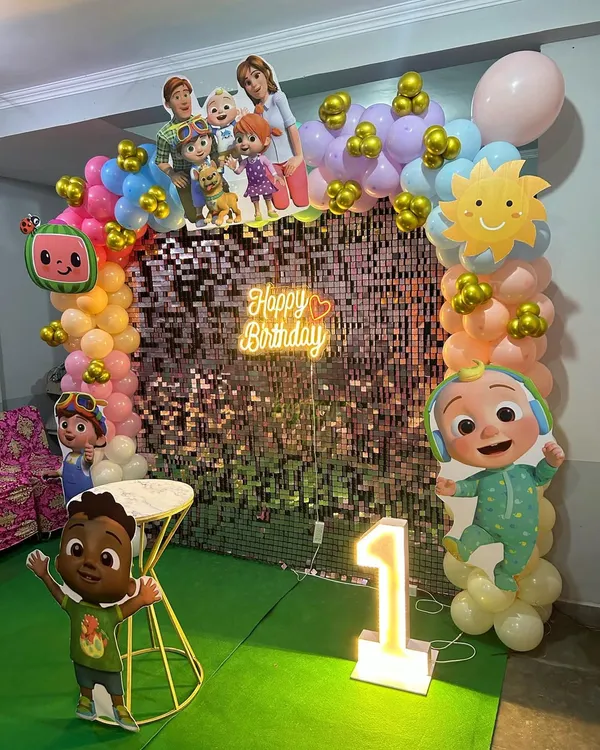 A vibrant Cocomelon theme first birthday party. This setup features a shimmering sequin backdrop, a 'Happy Birthday' neon sign, a colorful balloon arch, and multiple character cutouts from the popular show.