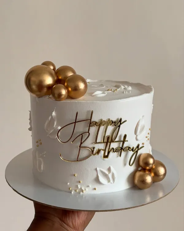 Simplicity at its finest. This all-white whipped cream cake is decorated with a "Happy Birthday" topper and delicate gold spheres for a clean, modern, and elegant look.